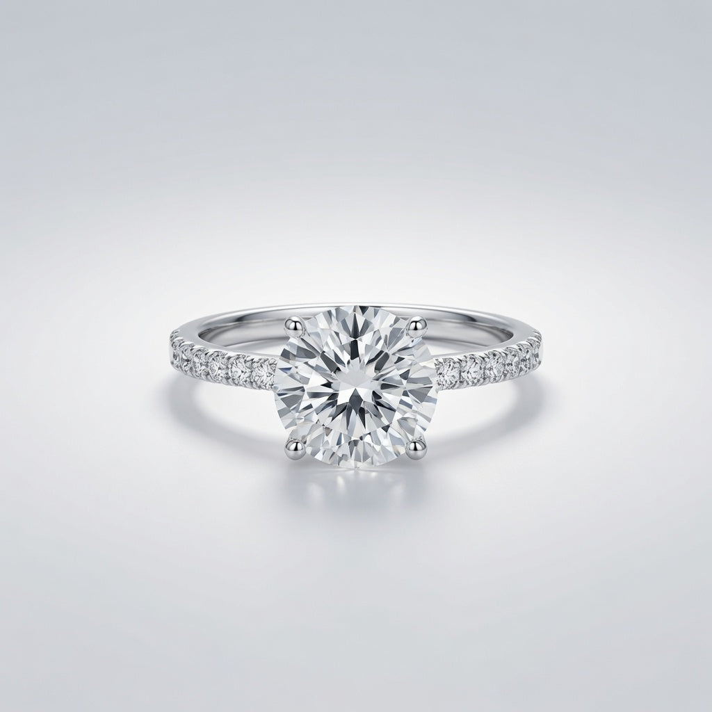 A classic solitaire engagement ring featuring a round brilliant-cut center diamond in a four-prong setting, with a pavé band set with smaller diamonds, shown on a light gray gradient background.