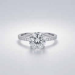 A classic solitaire engagement ring featuring a round brilliant-cut center diamond in a four-prong setting, with a pavé band set with smaller diamonds, shown on a light gray gradient background.